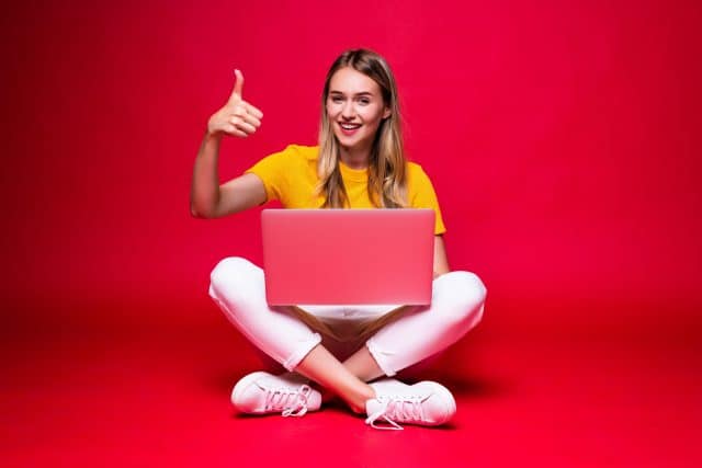 happy-young-curly-beautiful-woman-sitting-floor-with-crossed-legs-using-laptop-red-wall